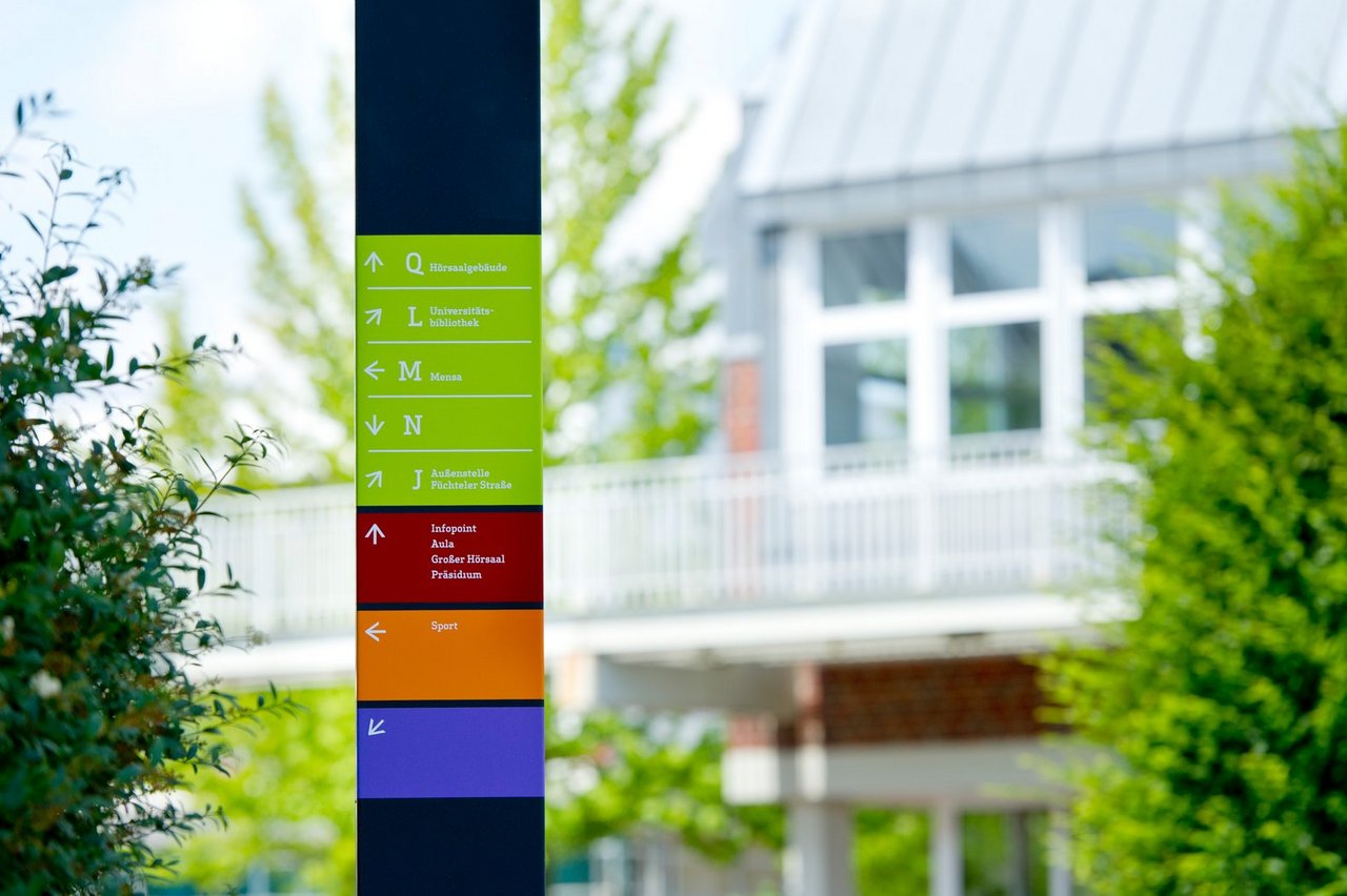 In the foreground, there is a guidepost of the campus guidance system. In the background, there is the bridge and the University Library (photo: University of Vechta/Meckel). 