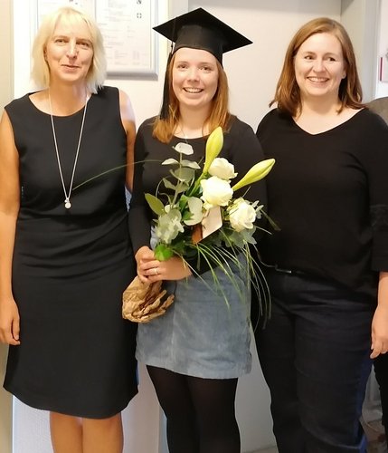 Ilka Gummels steht mit einem Blumenstrauß in der Hand und einem gebastelten Doktorhut auf dem Kopf zwischen ihren Doktormüttern.