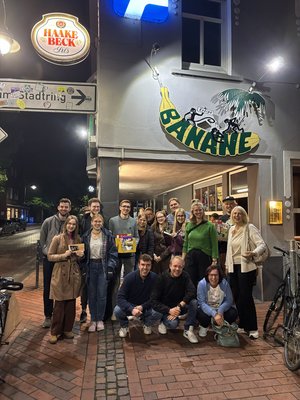 Das Foto zeigt die Gruppen, die beim Kneipenquiz die ersten drei Plätze belegt haben. Die Personengruppe steht draußen vor dem Gebäude der Banane unterhalb des Schriftzugs Banane.