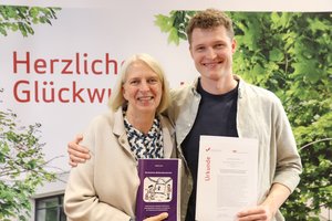 Zwei Personen stehen nebeneinander vor einer Wand auf der ein Teil des Schriftzugs 'Herzlichen Glückwunsch' zu lesen ist. Professorin Doktorin Martina Döhrmann steht links von Gerrit Loth. Frau Döhrmann hält das Buch zur Doktorarbeit von Gerrit Loth in der Hand und Gerrit Loth hält seine Urkunde in der Hand.
