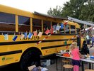 Seitenansicht eines amerikanischen Schulbus davor sieht man Kinder an einem Tisch mit Druck- und Bastelutensilien. 