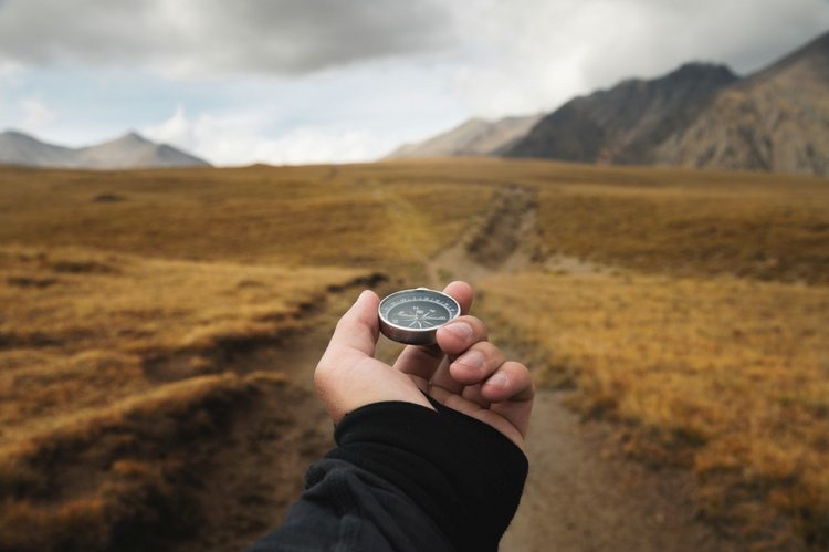 Die Hand eines Mannes, die einen Magnetkompass hält, in der Ich-Perspektive vor dem Hintergrund eines Höhenwegs und von Bergen