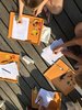 Kinder sitzen am Tisch und füllen etwas schriftlich aus. Man sieht Tablets mit ausgeschnittenen Figuren vor den Kindern.und Bastelutensilien auf dem Tisch.