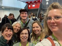 Auf dem Foto ist eine Gruppe von sechs Personen zu sehen, das in Form eines Selfies aufgenommen wurde. Die Personengruppe ist in einem großen Foyer mit vielen anderen Personen. 