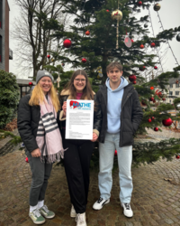 Vor einem geschmückten Weihnachtsbaum stehen zwei Studentinnen und ein Student. Die Studentin, die in der Mitte steht hält ein Dokument von 'Mathe im Advent' in der Hand auf der die eingereichte Aufgabe abgebildet ist.