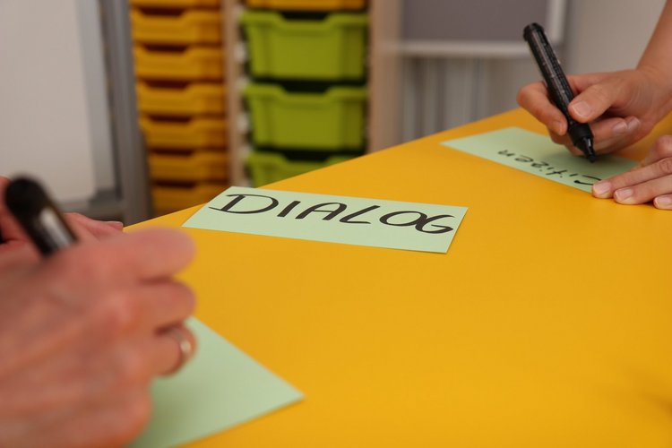 Moderationskarte Dialog. Foto: Friedrich Schmidt Zwei Personen schreiben auf Moderationskarten. Auf einer Karte in der Mitte steht Dialog geschrieben.