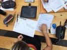 Aufsicht auf einen Arbeitsplatz einer Teilnehmerin. Ipad, Sifte, Aufgabenblatt und weitere Dinge befinden sich um sie herum. 