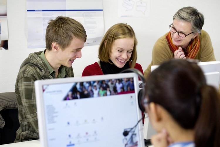 Zwei Studierende sitzen vor einem Computer. Sie unterhalten sich dabei ausgelassen mit einer Lehrenden.