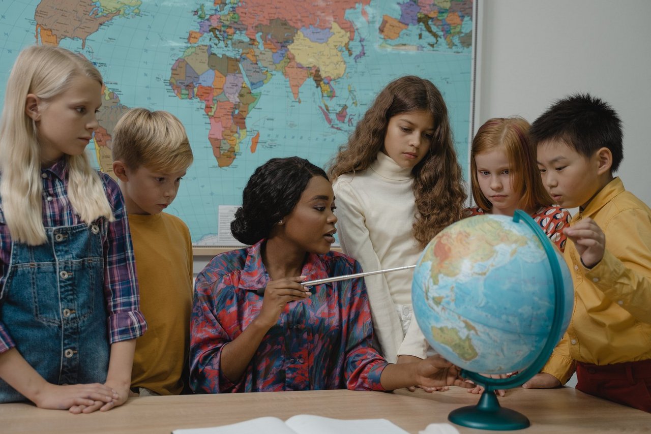 Lehrerin erklärt Schüler*innen etwas an einem Globus. Im Hintergrund hängt eine Weltkarte.