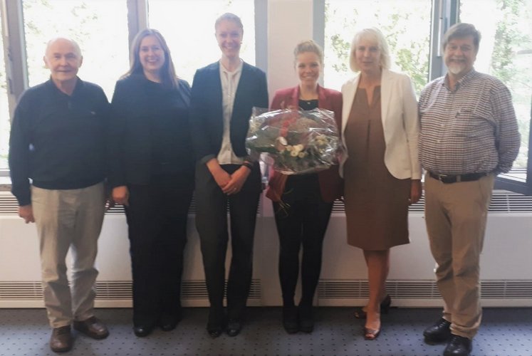 Sechs Personen stehen vor einem Fenster. Die zweite Person von rechts hält einen Blumenstrauß in den Händen.