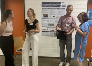 Vier Personen stehen in einem Raum mit hellem Teppichboden und einer weißen Ziegelwand. Im Hintergrund hängt ein wissenschaftliches Poster mit dem Titel „Coden von Zahlenfolgen für Anfänger*innen: Ein fächerverbindendes Konzept für Lernende und Lehrkräfte im Projekt CoM-MIT“.Auf der linken Seite unterhalten sich zwei Frauen im Profil miteinander und lachen. Die linke Frau trägt ein helles Top und eine dunkle Hose, die rechte Frau ein schwarzes Oberteil und eine weite, weiße Hose.Auf der rechten Seite stehen ein Mann und eine Frau. Der Mann trägt ein rot-weiß kariertes Hemd, eine graue Jeans und hält ein aufgeklapptes, dunkles Tablet in den Händen. Er blickt lächelnd zu der Frau rechts neben ihm, die ein hellblaues Kleid und eine Brille trägt. Beide Gruppen tragen Namensschilder. Im Hintergrund sind zwei orangefarbene Türrahmen und zwei Holzstühle zu sehen.