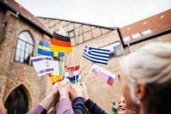 Das Bild zeigt eine Studierendengruppe, die verschiedene EU Länderflaggen nach oben zusammenhalten.
