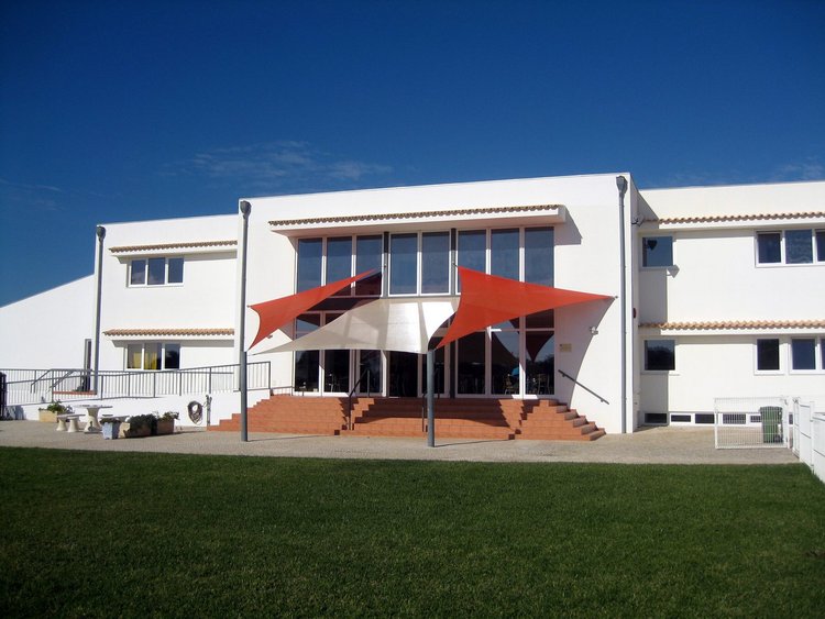 Eingang der DS Algarve. Zu sehen ist ein weißes Gebäude mit roten Sonnensegeln vor der Eingangstür.