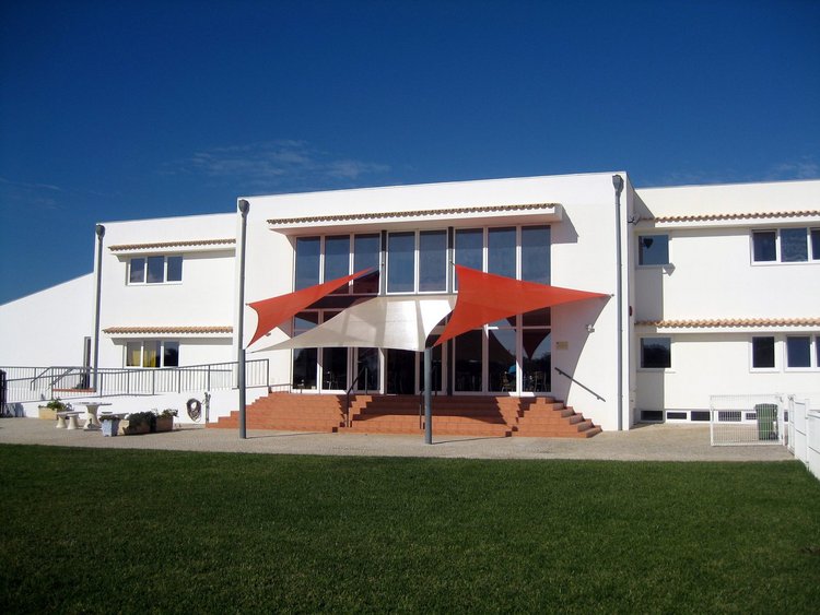 DS Algarve Eingang der DS Algarve. Zu sehen ist ein weißes Gebäude mit roten Sonnensegeln vor der Eingangstür.