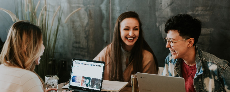 Drei Studierende sitzen mit Laptops zusammen an einem Tisch und lachen.